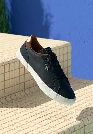 Schwarze Leder-Sneaker mit weißer Sohle und braunen Akzenten, die auf beigen Fliesenstufen vor einer blauen Wand präsentiert werden.