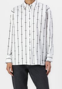 Chemise blanche boutonnée avec un motif à rayures noires et motifs graphiques. Le tissu est lisse avec des manches longues et des poignets boutonnés.