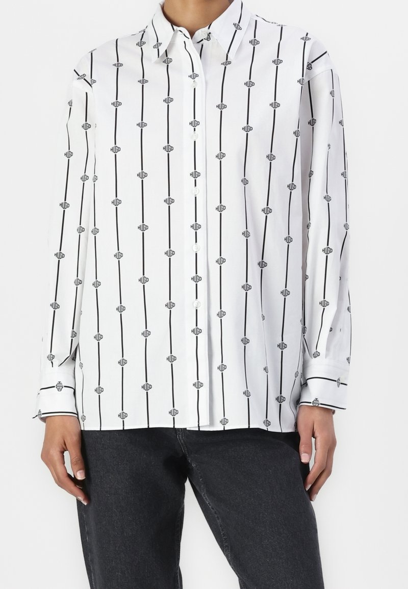 Chemise blanche boutonnée avec un motif à rayures noires et motifs graphiques. Le tissu est lisse avec des manches longues et des poignets boutonnés.