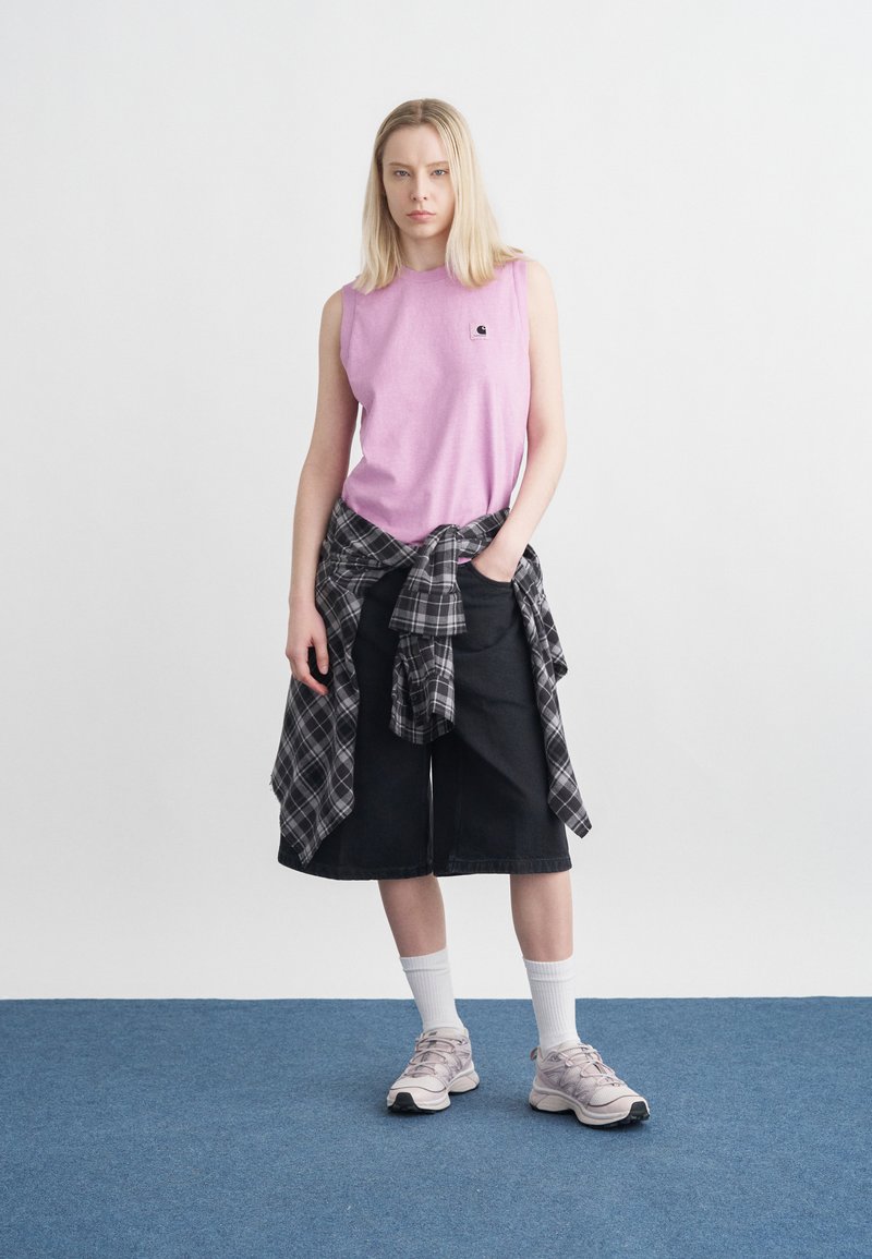 Young woman with blonde hair wearing sleeveless pink top, black knee-length shorts, tied plaid shirt, white socks, and pink sneakers, standing on blue carpet.