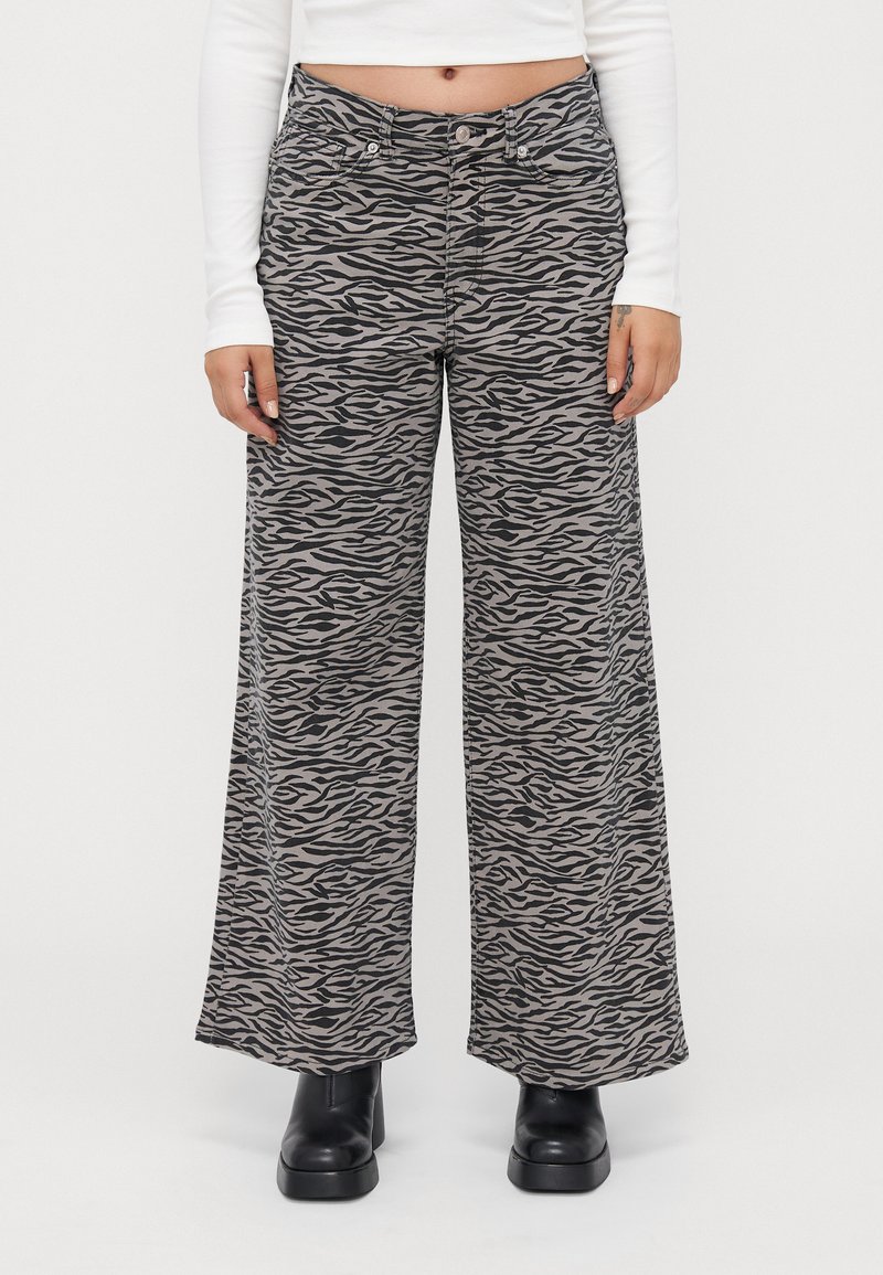 Mujer vistiendo pantalones de pierna ancha con estampado de cebra en gris y negro, top corto de manga larga blanca y botas de tobillo negras con plataforma.