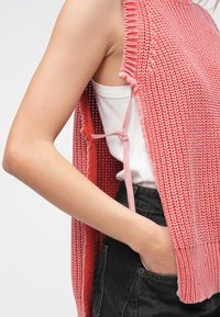 Πλεκτό αμάνικο τοπ με χαλαρή εφαρμογή, σε αποχρώσεις του ροζ. Διαθέτει διαπλεκόμενη υφή, λεπτομέρεια με δετή πλευρά και φοριέται πάνω από λευκό φανελάκι.