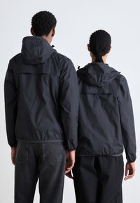 Zwarte lichtgewicht jassen hebben capuchons en elastische manchetten, weergegeven op twee figuren met een gladde textuur, gecombineerd met donkere spijkerbroeken.