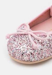 Bailarina brilhante em rosa com parte superior macia, apresentando um detalhe de laço e uma superfície texturizada. A sola é plana e de um bege claro.