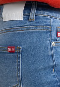 Zili džinsi ar gaismu mazgāšanu, ar sarkaniem logo tagiem aizmugurējos kabatās, kontrastējošiem dūriem un baltu jostas birku.