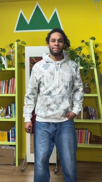 Camisola com capuz cinza claro com padrão de folhas e logótipo "GAP", combinada com jeans de ganga azul. O fundo apresenta paredes amarelas e prateleiras verdes.