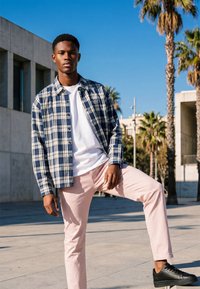 Jeune homme portant une chemise à carreaux bleue, un t-shirt blanc, un pantalon rose clair et des baskets noires, debout à l’extérieur avec un pied posé sur un rebord, des palmiers en arrière-plan.
