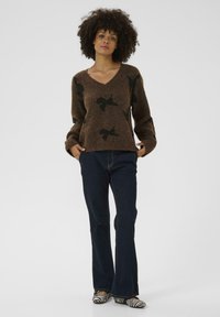 Brauner Strickpullover mit schwarzem Blumenmuster, V-Ausschnitt und langen Ärmeln. Kombiniert mit dunkelblauen Schlagjeans und Schuhen im Zebra-Print.