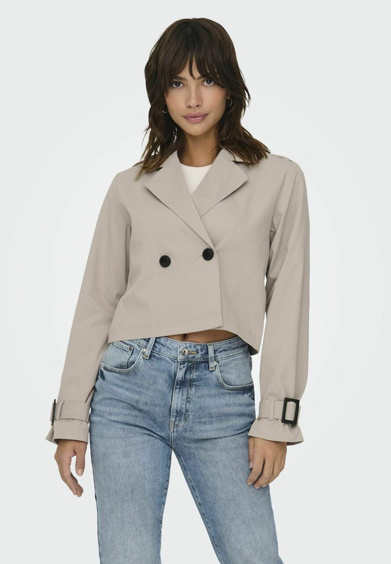 Veste beige cintrée avec des boutons croisés, de larges revers et des sangles aux poignets. Portée sur un top blanc et associée à un jean bleu clair.