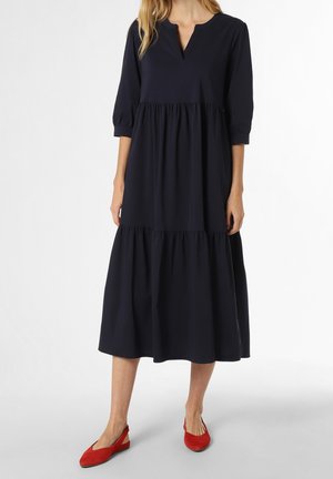 Frau trägt ein locker sitzendes, langes schwarzes Kleid mit dreiviertel Ärmeln und roten flachen Schuhen, steht auf einem schlichten hellen Hintergrund.