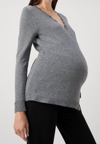 Kobieta w ciąży w dopasowanym, szarym swetrze i czarnych spodniach, pokazana od szyi do bioder na jednolitym tle.