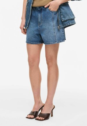 Kvinne iført blå denimshorts med høyt liv, brune teksturerte sandaler med hæl og en blå denimjakke med stor lomme og glidelåsdetalj.