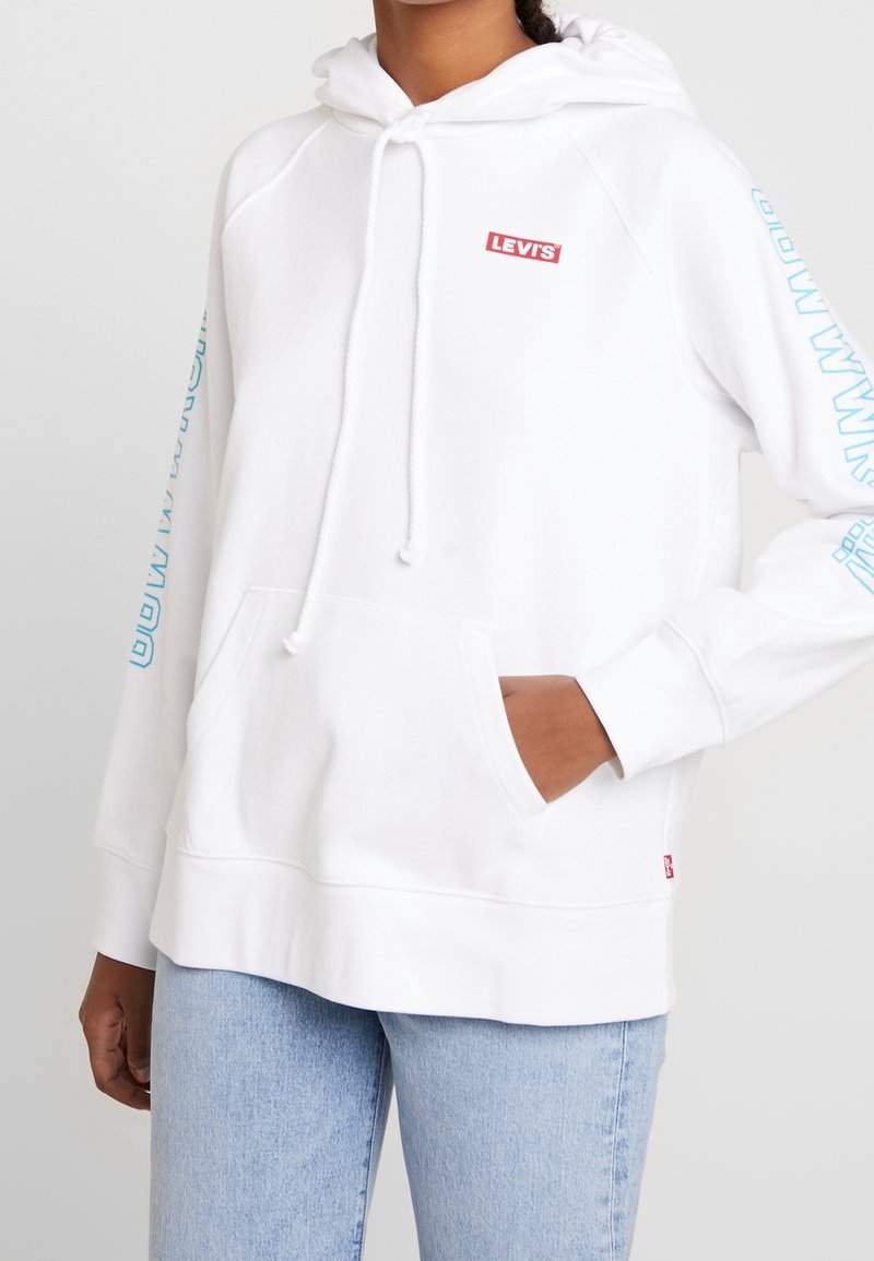 Sweat à capuche blanc en mélange de coton, doté d'une poche avant, d'une capuche avec cordon de serrage, et d'un logo Levis rouge avec du texte bleu sur les manches.