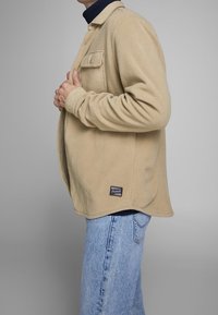 Beżowa kurtka koszulowa z polaru z zapięciem na guziki, jedną kieszenią na piersi i lekko zaokrąglonym dołem. Połączona z jasnoniebieskimi dżinsami.