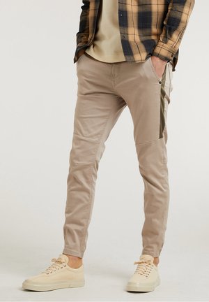 Pantalon beige ajusté, avec poches latérales et accents verts contrastants. Fabriqué en tissu doux, associé à des baskets de couleur claire.