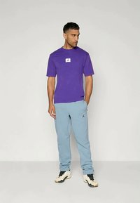 Cămașă scurtă violet cu un patch alb cu logo, asociată cu pantaloni scurți de trening bleu deschis cu un mic logo negru, purtată cu încălțăminte alb-negru.