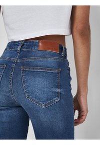 Jeans de denim azul oscuro con un diseño ajustado, con una etiqueta de cuero marrón que dice "NOISY MAY" en la cintura trasera.