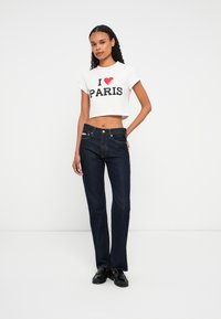 Femeie îmbrăcată cu un tricou alb scurt cu textul "I ♥ PARIS", blugi negri și mocasini negri, stând în fața unui fundal alb.
