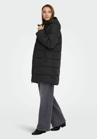 Melna polsterēta jaka ar kapuci, ar rāvējslēdzēja aizdari un sānu kabatām. Uzvilkta virs platām pelēkajām džinsām un potīšu zābakiem, izrādīta uz modele.