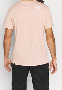 Růžové bavlněné tričko s krátkými rukávy, kulatým výstřihem a bílým logem na horní části zad. Nosené s černými kalhotami.