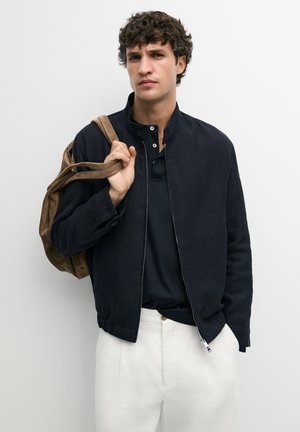 Jeune homme aux cheveux bouclés portant une veste noire et un pantalon blanc, tenant un sac à dos marron sur son épaule sur un fond uni.