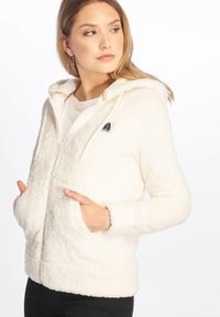 Sudadera con cremallera de color blanco sucio y textura suave, con capucha ajustable, bolsillos frontales y un pequeño parche con el logo en el pecho. Tejido suave y texturizado.