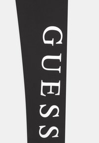 Zwarte legging met het witte "GUESS" logo verticaal gedrukt, met een gladde textuur en rekbare stof voor flexibiliteit.