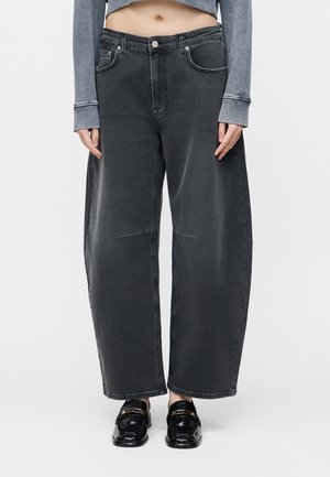 Άτομο που φορά φαρδιά, ξεθωριασμένα μαύρα τζιν, γκρι cropped τοπ με μακρύ μανίκι και μαύρα δερμάτινα loafers με χρυσά αγκράφες.