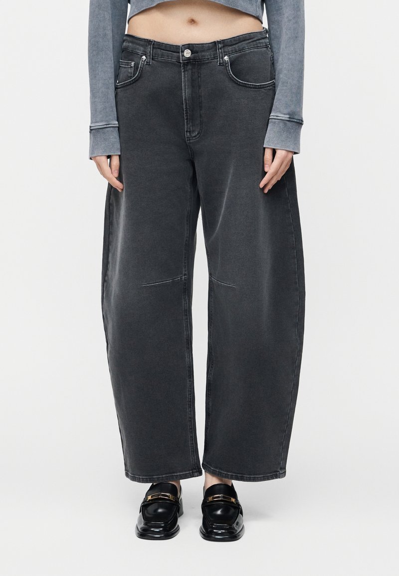 Άτομο που φορά φαρδιά, ξεθωριασμένα μαύρα τζιν, γκρι cropped τοπ με μακρύ μανίκι και μαύρα δερμάτινα loafers με χρυσά αγκράφες.