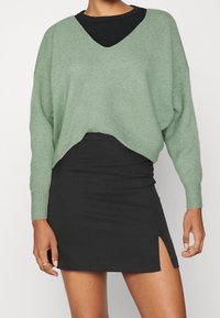 Maglione verde lavorato a maglia con scollatura a V, vestibilità ampia e polsini a coste, abbinato a una mini gonna nera con uno spacco laterale.