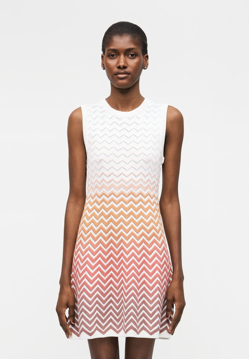 Γυναίκα φορώντας αμάνικο πλεκτό φόρεμα με λευκό, πορτοκαλί και ροζ ζικ-ζακ μοτίβο διαβάθμισης, στέκεται μπροστά από απλό φωτεινό φόντο.