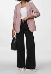 Blazer rose clair porté sur un haut blanc, associé à un jean noir large taille haute et des baskets noires, avec un petit sac à main noir.