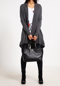Cardigan gris à devant ouvert et forme fluide, porté sur un haut graphique blanc. Sac à main en cuir noir et pantalon sombre ajusté avec bottines noires.