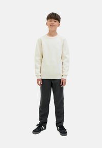 Light cream, textured sweater with ribbed cuffs, paired with dark gray patterned pants and black sneakers. Model stands with hands relaxed.