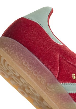 Zapatillas de terciopelo rojo con acentos en verde menta y una suela de goma transparente. Presentan un logotipo en relieve y detalles de costura distintivos.