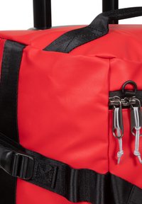Červená látková sportovní taška s černými popruhy, obsahující zipy a kovové komponenty. Texturovaný povrch s nastavitelnými sponami a elastickými šňůrami.