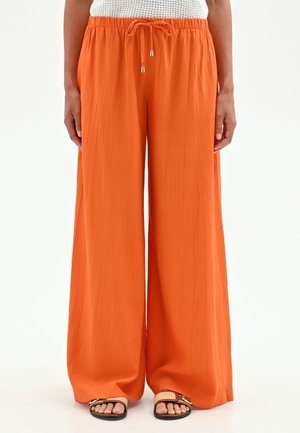 Pantaloni larghi a gamba larga di colore arancione brillante con vita elasticizzata e coulisse, indossati con sandali aperti.