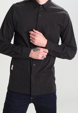 Mężczyzna w ciemnoszarej koszuli zapinanej na guziki, z tatuażami na dłoniach i palcach, poprawiający lewy rękaw na jednolitym jasnym tle.