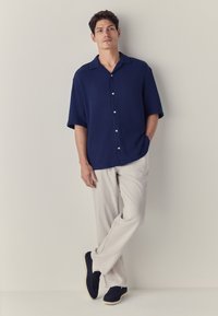 Tamnoplava muška košulja kratkih rukava s gumbima i teksturiranim tkanjem, uparena sa svijetlobéž hlačama i tamnim cipelama. Jednostavan, opušten kroj.