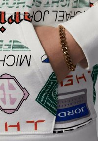 Felpa bianca con testo e grafiche colorate, con un primo piano di un braccialetto in catena dorato su un polso.