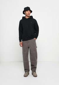 Felpa nera con cappuccio e lacci, pantaloni grigi larghi con cuciture visibili e stivali grigi. Il modello indossa un cappello a secchiello nero.