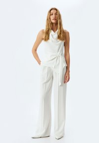 Top bianco senza maniche con scollo a cowl e vita legata. Abbinato a pantaloni bianchi a gamba larga e tacchi a punta. Tessuto morbido, design minimale.