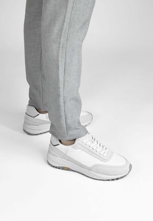 Persoon die lichtgrijze broek en witte sneakers met een gestructureerde zool draagt, staand op een witte ondergrond.