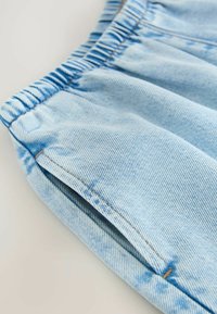 Detaliu pantaloni din denim albastru deschis care arată talie elastică și buzunar lateral cu cusături vizibile.