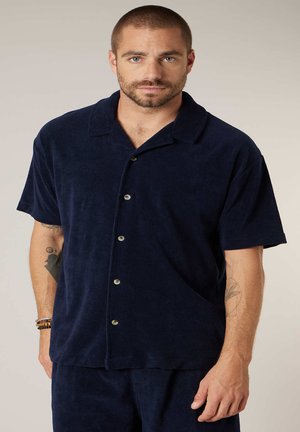 Man met kort haar en baard, gekleed in een marineblauw overhemd met korte mouwen en bijpassende broek, met tatoeages op beide armen zichtbaar.