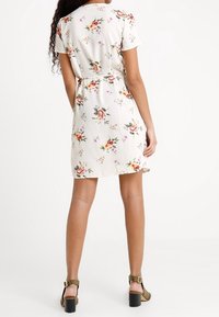 Květinové šaty s bílým základem, krátkými rukávy a páskem v oblasti pasu. Obsahují vícoloré květinové vzory a mírně nadkolenní délku.