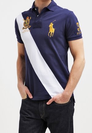 Polo azul marino con una franja diagonal blanca, logo amarillo bordado en el pecho y tapeta con tres botones. Tela de algodón, mangas cortas.