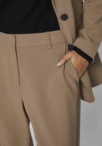 Beige skræddersyede bukser med en glat tekstur, der har sidelommer og en frontfold. Modelens hånd hviler i lommen.