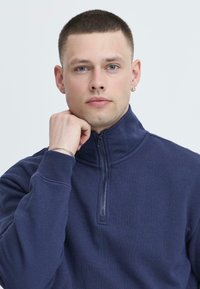 Felpa zip-in blu navy con colletto alto, caratterizzata da un tessuto strutturato e polsini a coste. Il modello posa con la mano sul mento.