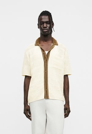 Männliches Model mit dunkler Haut und geflochtenem Haar trägt ein beige gestricktes, kurzärmeliges Hemd mit Knöpfen und brauner Borte sowie helle Hosen.